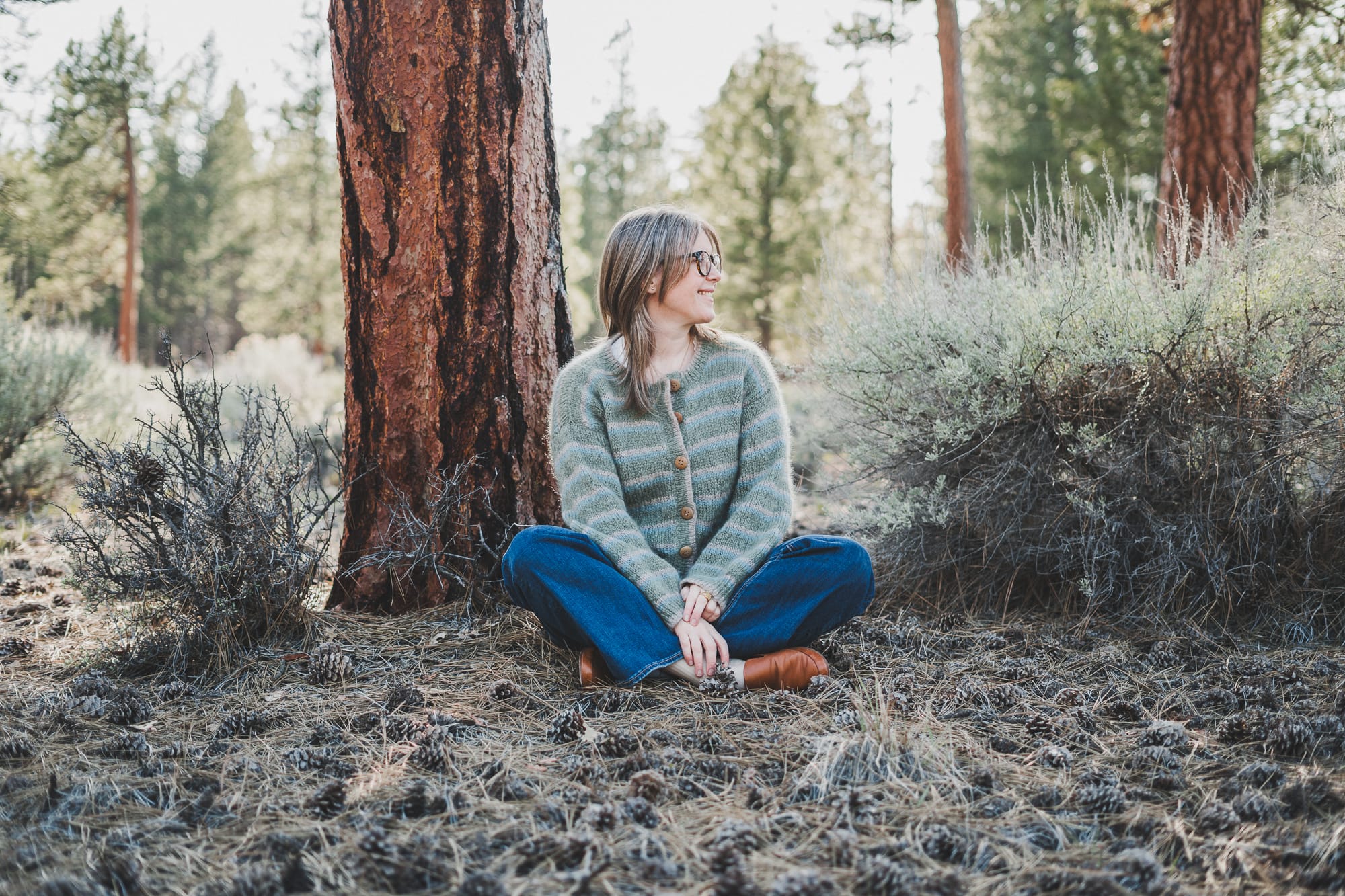 Chloé sitting down in the forest wearing her Wool and the Gang Piece of Me Cardigan in colors latte green and blue