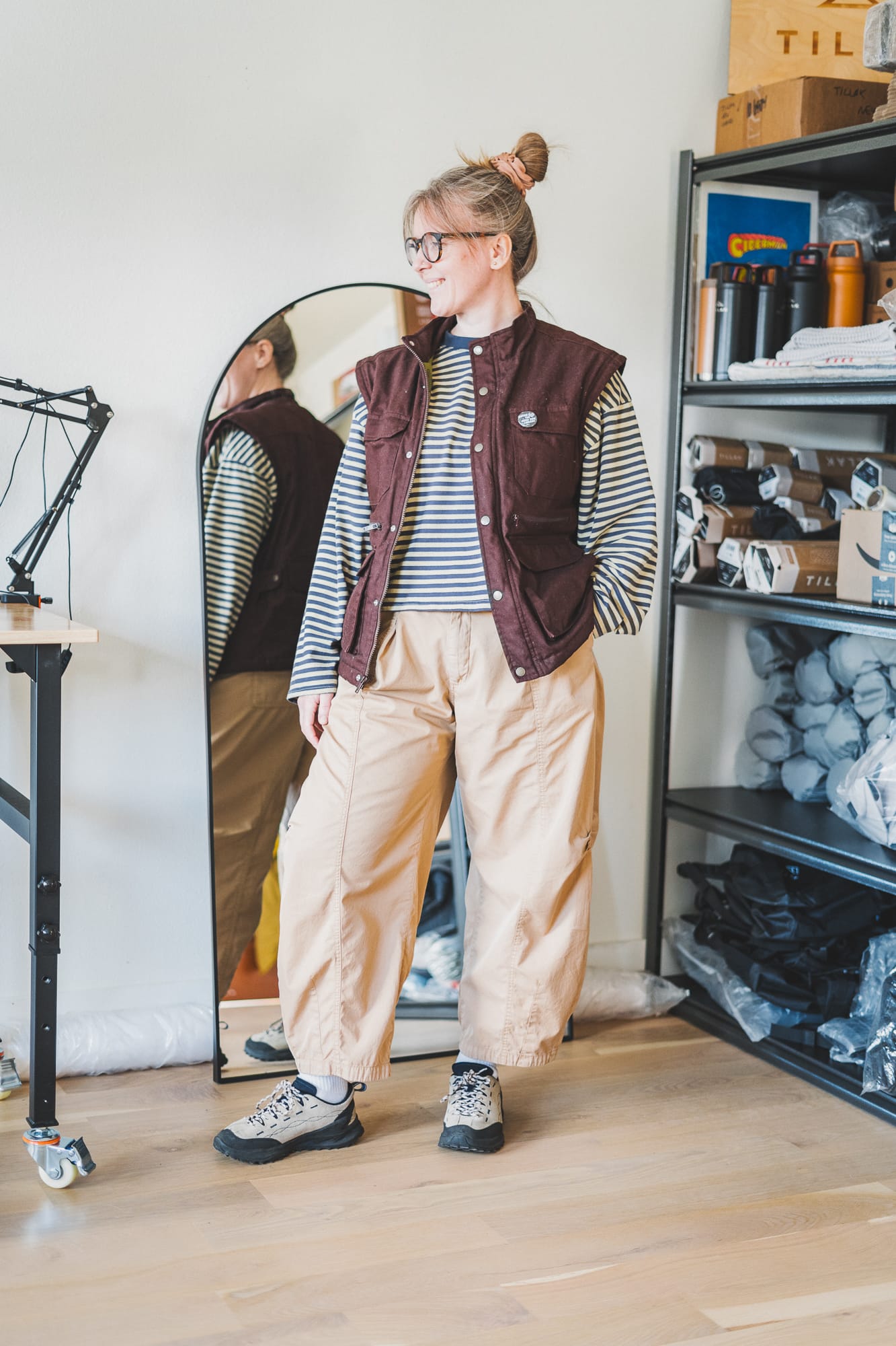 Person wearing tan barrel-leg pants, a striped long-sleeve shirt, and a burgundy utility vest, standing indoors beside a full-length mirror and shelving unit.
