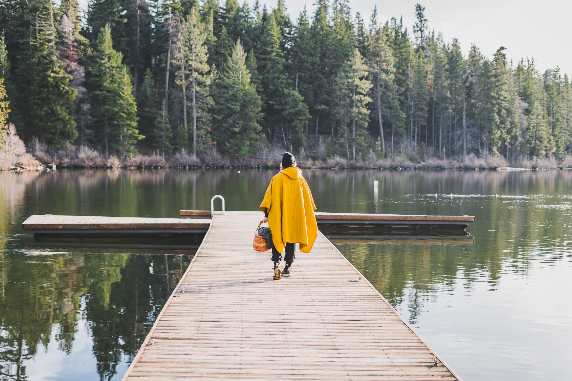 Person wearing a bright yellow poncho walks along a wooden dock toward a calm forest-lined lake, carrying a woven basket and bag, with evergreen trees reflected in the water.