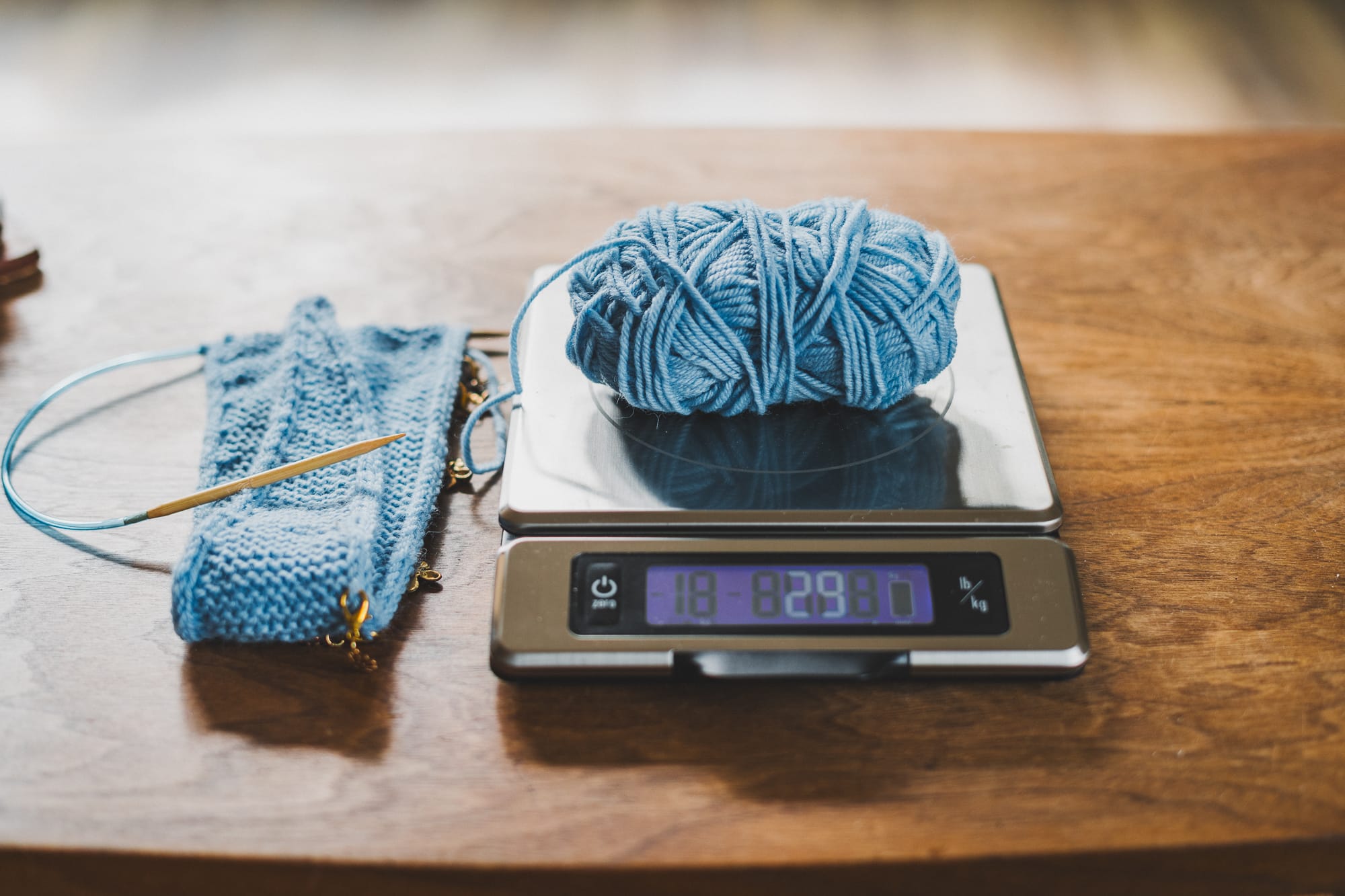 Light blue yarn ball on a kitchen scale next to a partially knitted Sophie scarf on circular needles, showing how to weigh yarn during a knitting project.