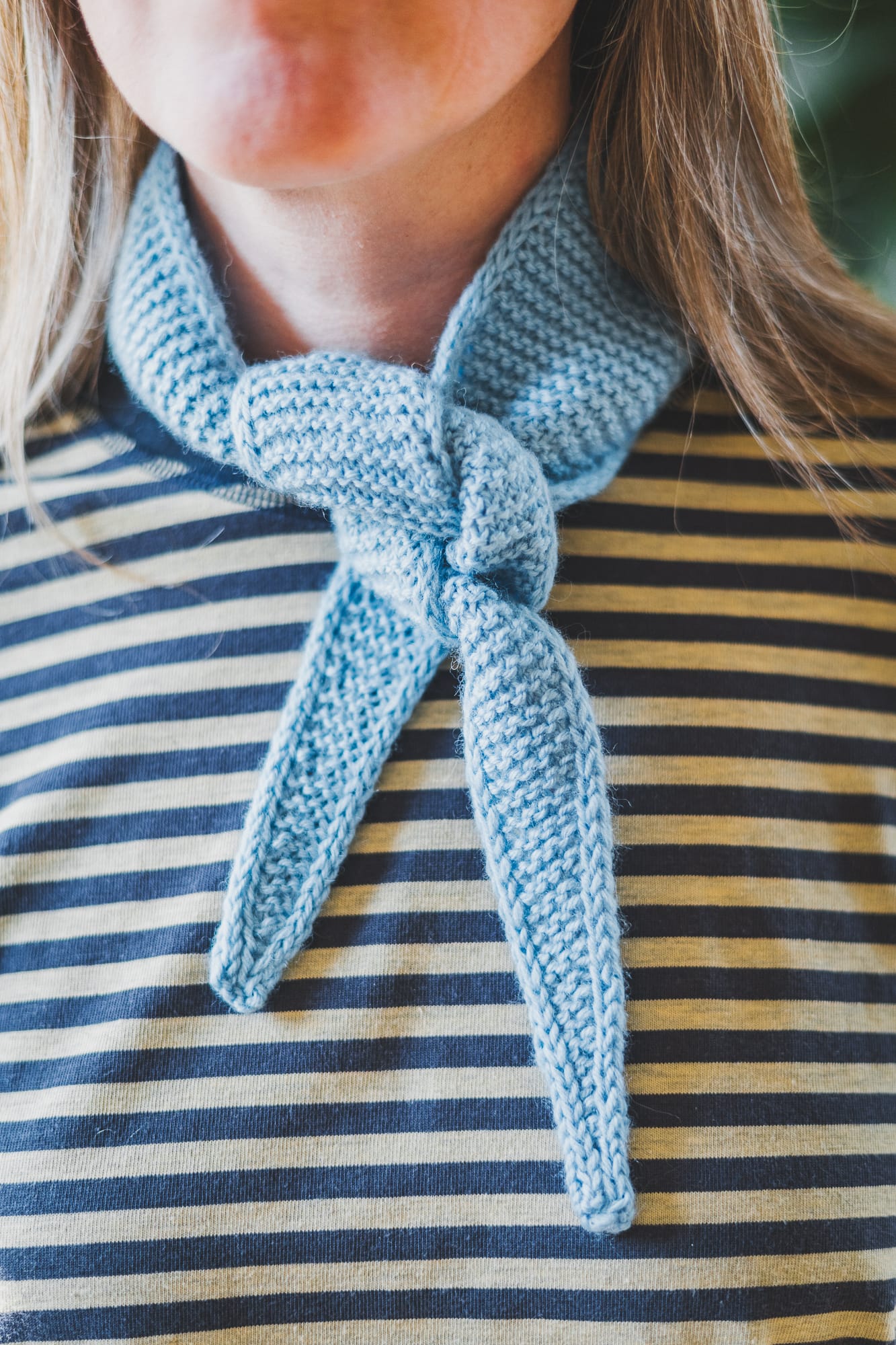 Close-up of a light blue hand-knitted Sophie Scarf by PetiteKnit tied at the neck, showing the garter stitch texture and i-cord edges against a striped shirt.