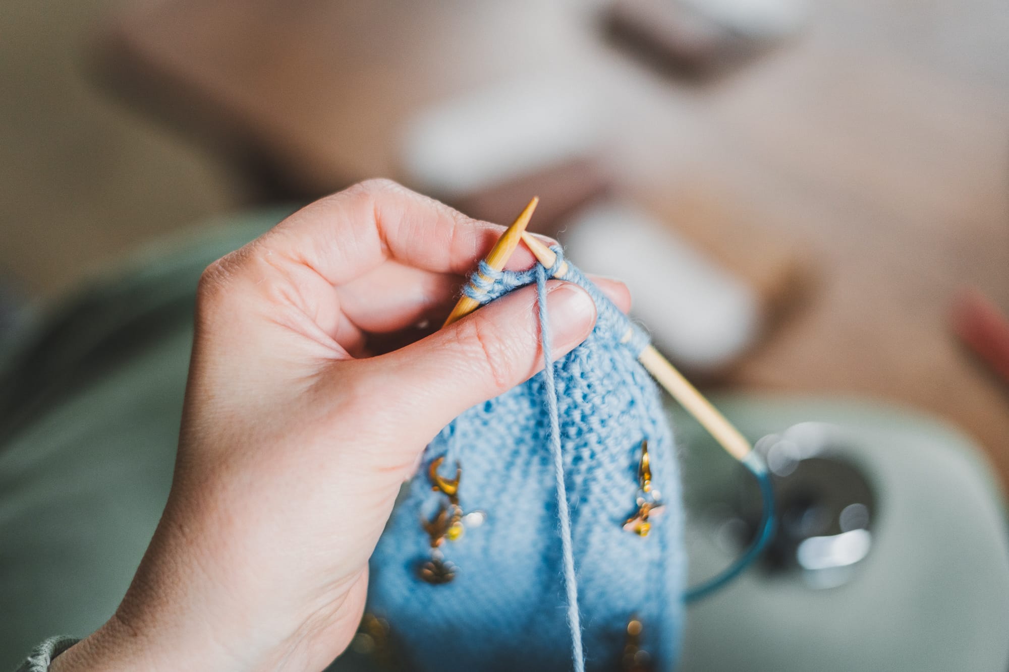Hands knitting a light blue Sophie scarf with circular needles and stitch markers, working on the i-cord edge while sitting at a table.